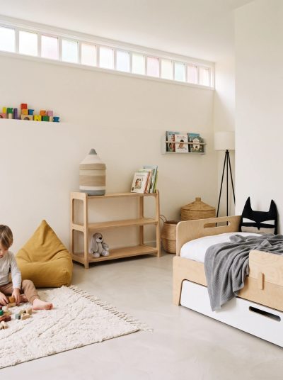 יחידה ישרה נמוכה של מדפי אחסון Corner מעץ ליבנה RANCO בחדר משחקים, עם סל, ספרים ובובה, ליד מיטת ילדים ופוף